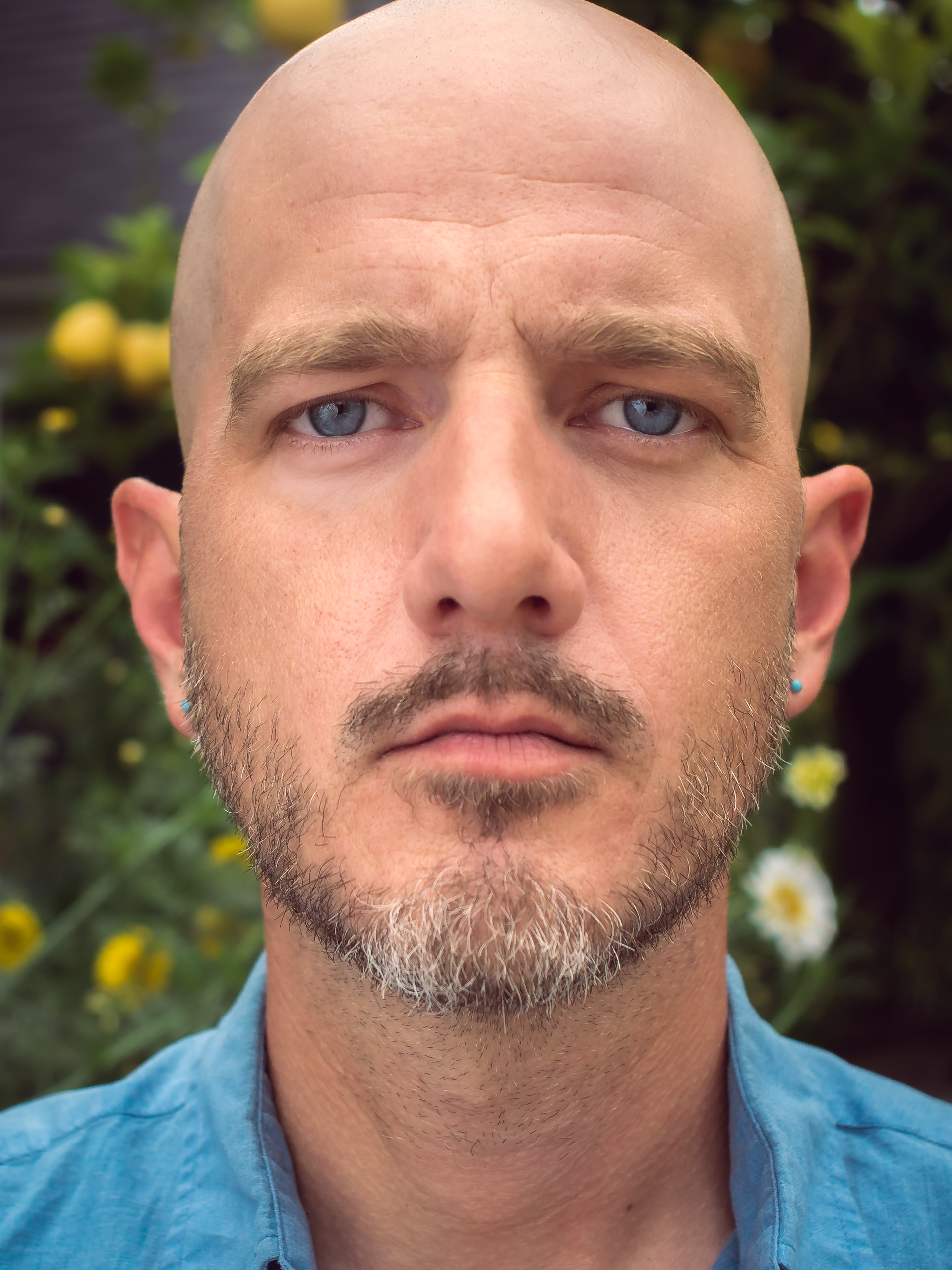 Self portrait of Los Angeles lifestyle and portrait photographer Ken Morris. It's a closely cropped image, Ken looks directly a the camera with a somber expression. He is bald with a short beard and Ken Morris wears a blue shirt. Flowers are behind him.